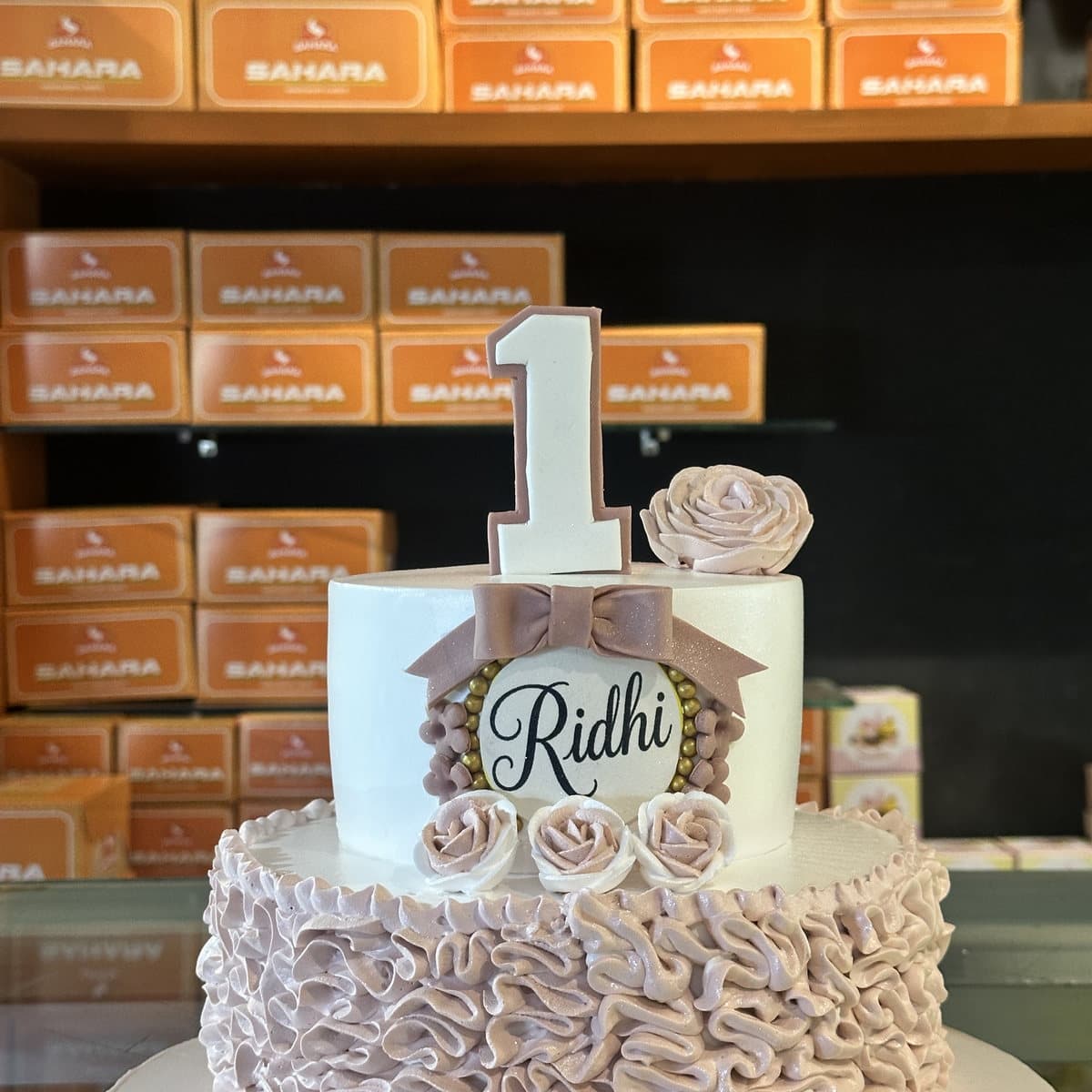 Two-tier first birthday cake with pink buttercream ruffles and a gold number one topper, made at Sahara Bakery Rewari