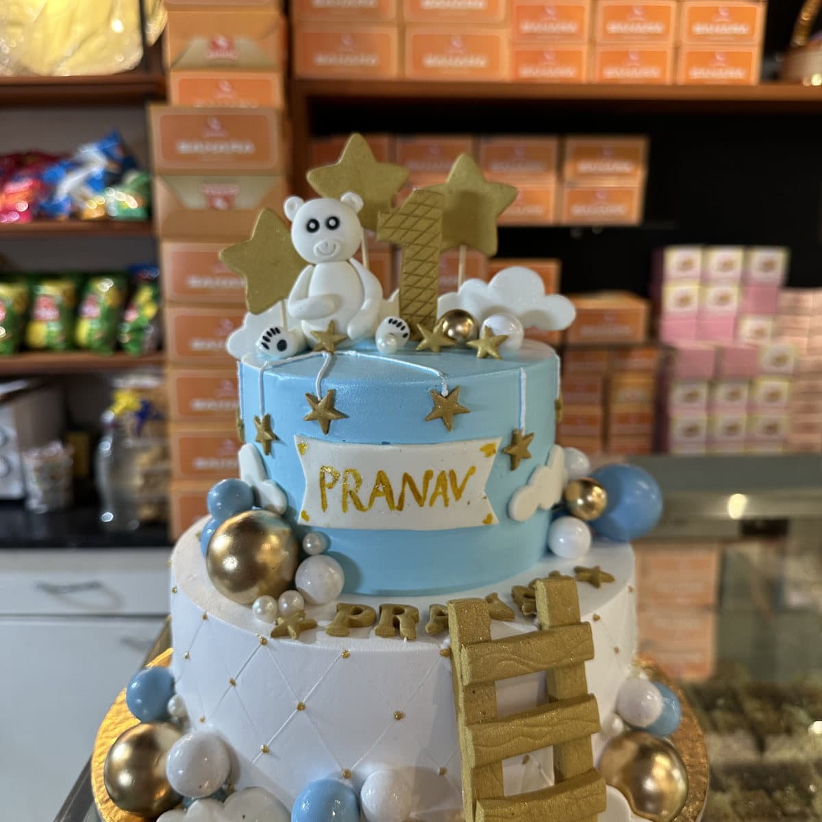 Two-tier blue and gold birthday cake with a teddy bear and cloud topper for a baby boy